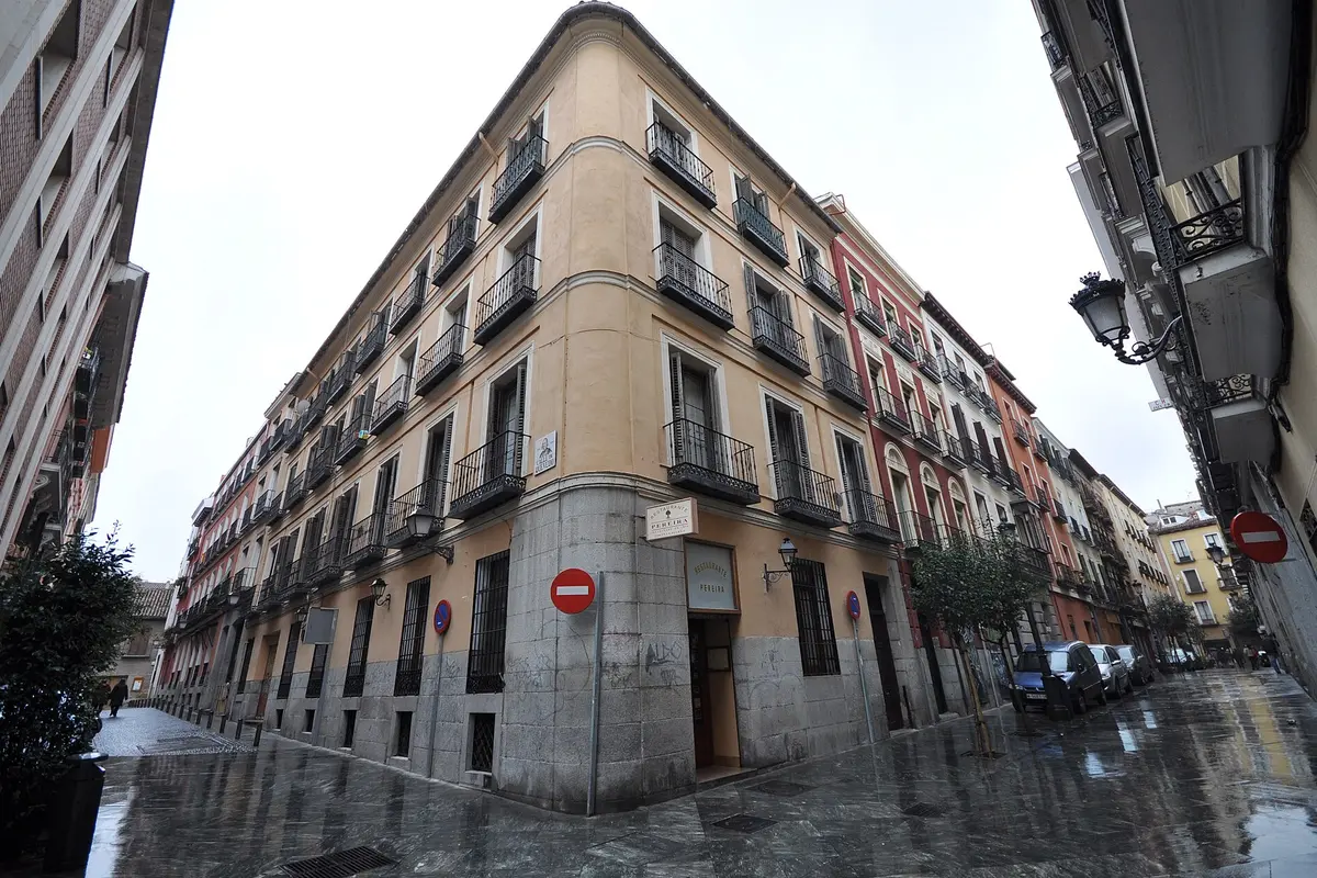 A wet pedestrian street in the Barrio de las Letras, Madrid, with an apricot-coloured corner building, iron balconies and cars parked along the street