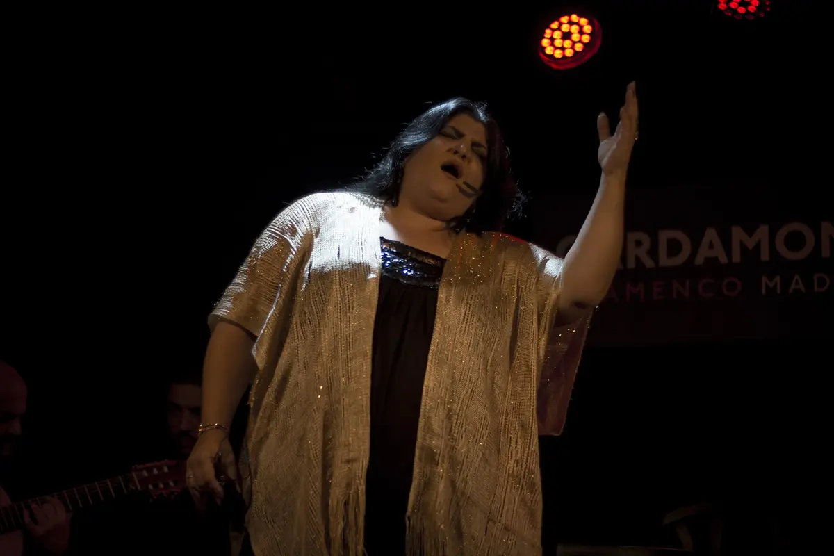 A singer on the stage at Cardamomo in Madrid, one arm raised, a guitarist partly visible at the left and the venue name glowing on the wall behind