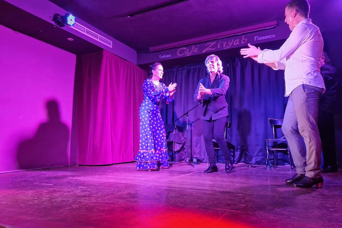 A dancer in a blue polka-dot dress, a guitarist and a second man clapping on a small stage under pink and blue lights